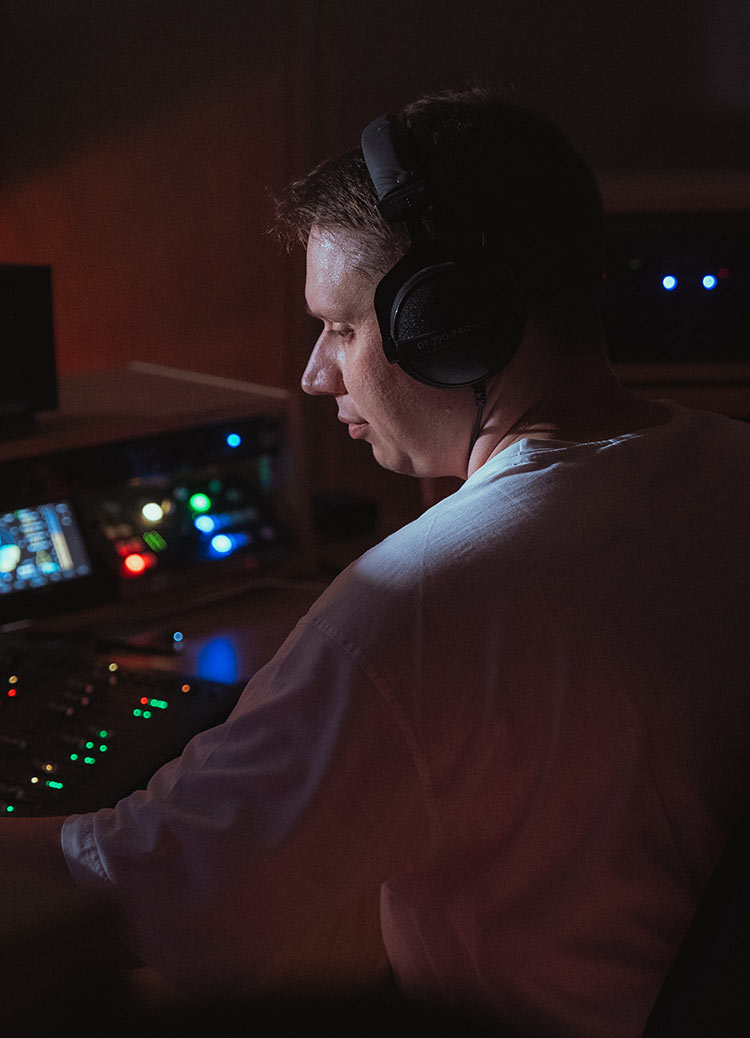 Jordi Büchel arbeitet mit aufgezogenen Beyerdynamic
DT 770 Pro Kopfhörern an einer Musikpostproduktion.
Das Licht des PC-Monitors erhellt sein Gesicht. Auf
dem Studio-Desk leuchten LEDs des darauf liegenden
Avid S3 Controller, des API MC530 Monitorcontroller
und des TC-Electronic Clarity M.